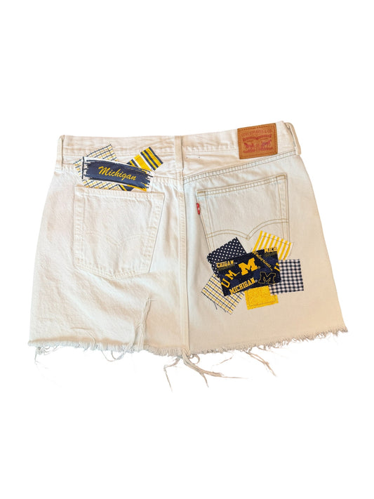 Off-white denim shorts with a distressed frayed hem, featuring a Michigan Wolverines patchwork design on the back pocket with navy, yellow, and white fabric pieces, and a brown leather patch on the waistband.