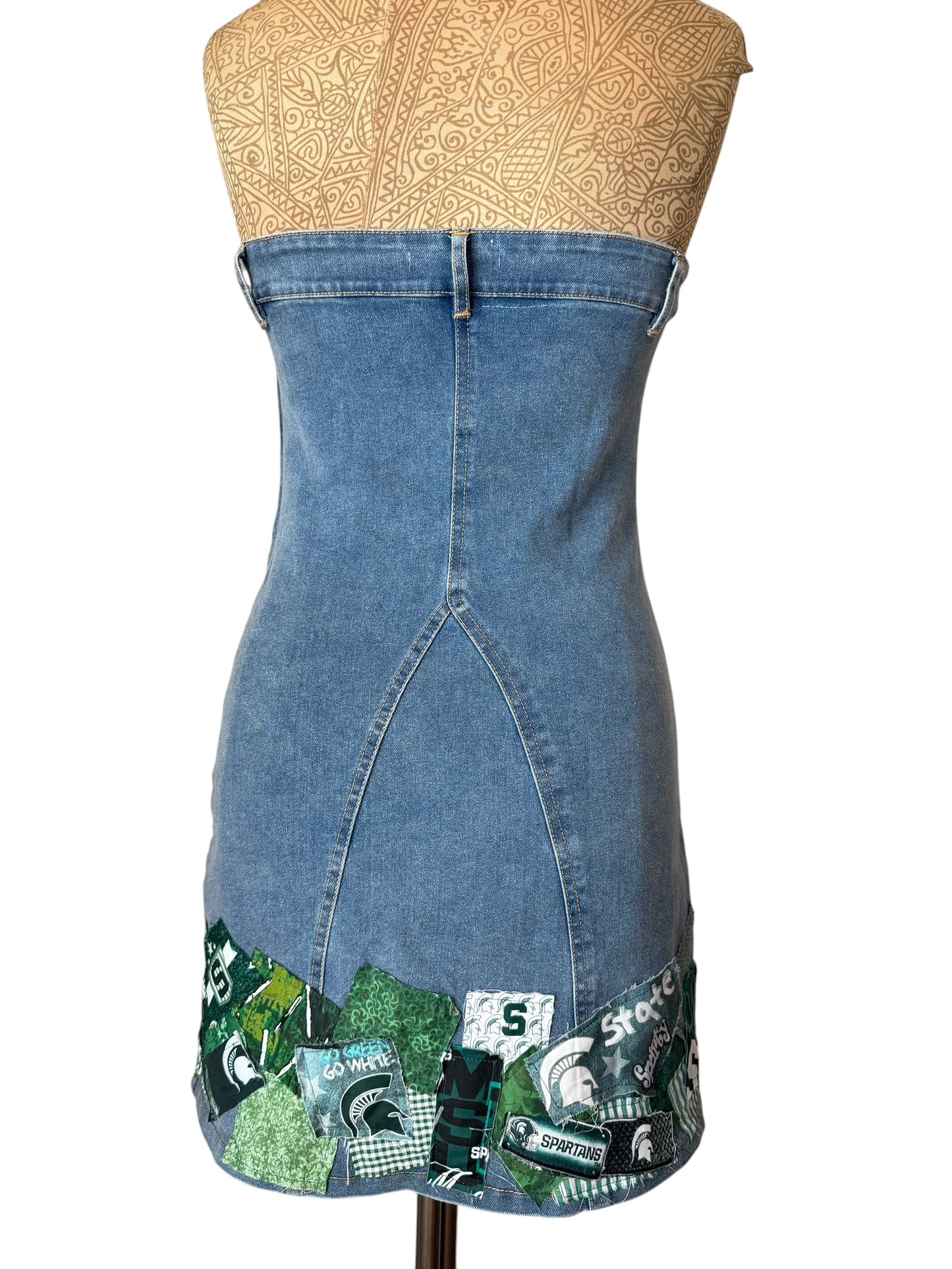 A strapless blue denim dress displayed on a tan mannequin torso against a white background, featuring a green and white patchwork trim at the hem with Michigan State Spartans-themed designs.