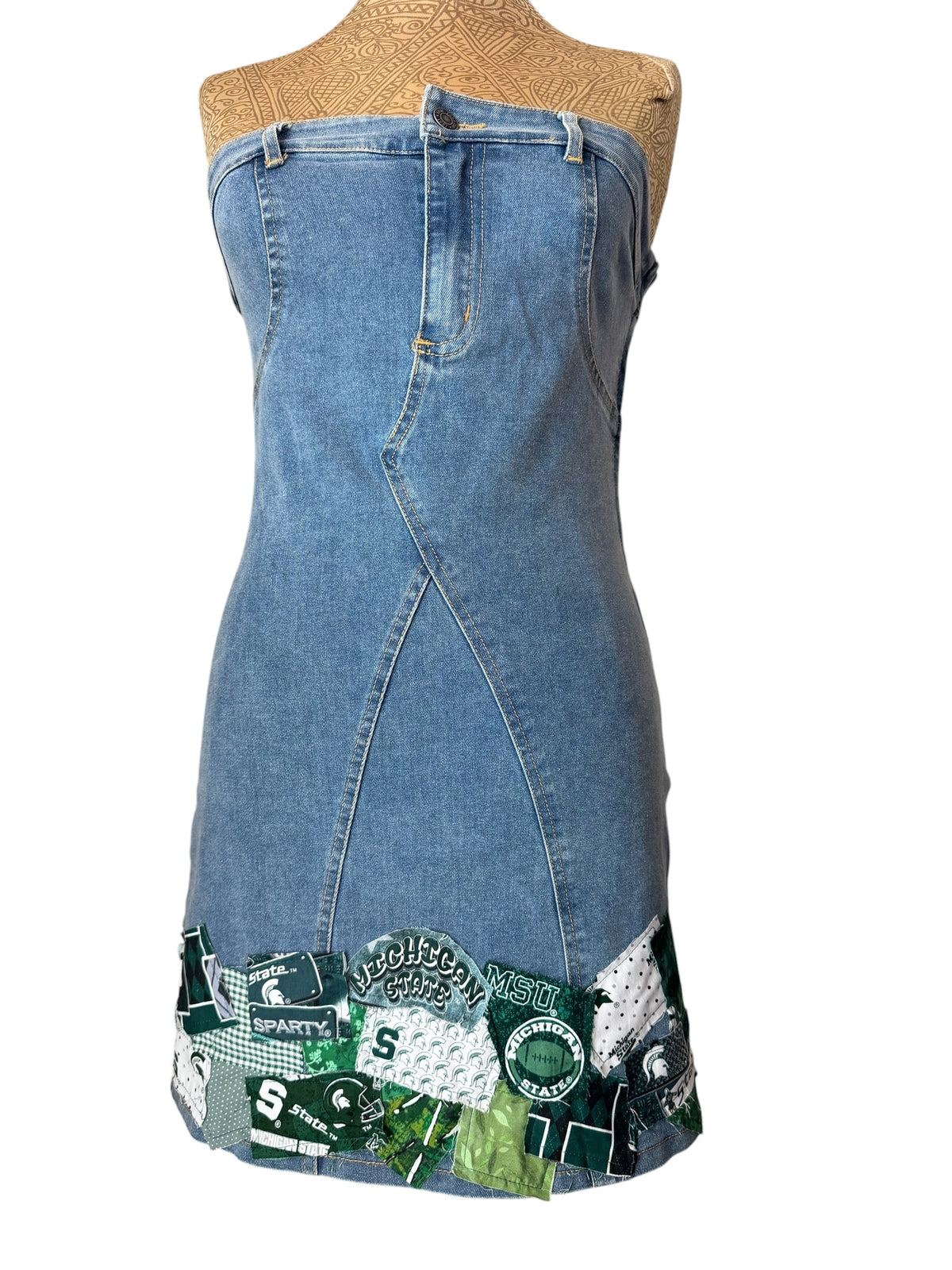 A strapless blue denim dress displayed on a tan mannequin torso against a white background, featuring a front wrap-style seam and a green collegiate-themed patchwork trim at the hem with visible text and logos.