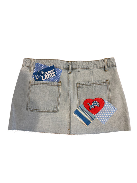A light gray denim mini skirt shown from the back, featuring two pockets with Detroit Lions themed patches, including a red heart patch and blue polka dot patches.