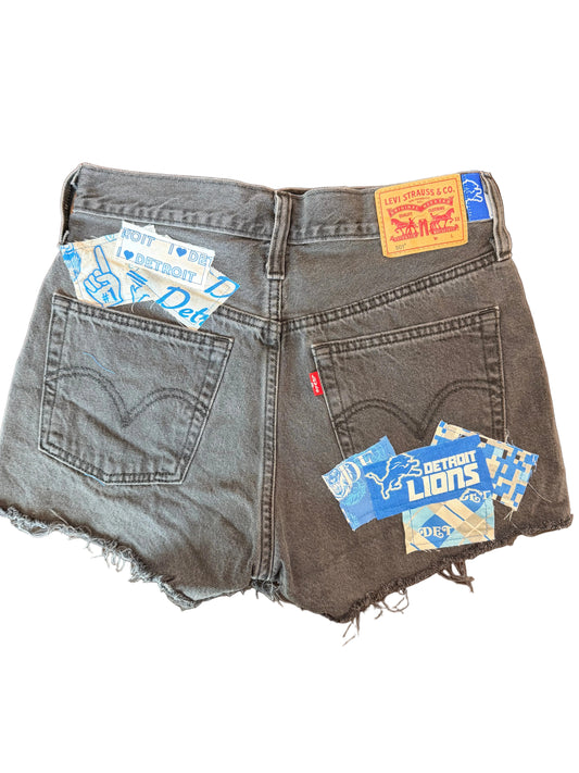 A pair of dark gray denim shorts with raw, frayed hems, featuring multiple colorful Levi's brand patches and labels attached to the back pockets, laid flat on a dark wooden surface.