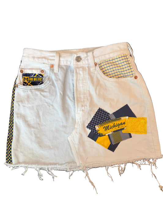 A pair of white denim shorts with a distressed frayed hem, featuring a Michigan Wolverines themed patchwork design with navy, yellow, and white patterned fabric pieces on the front pocket area.