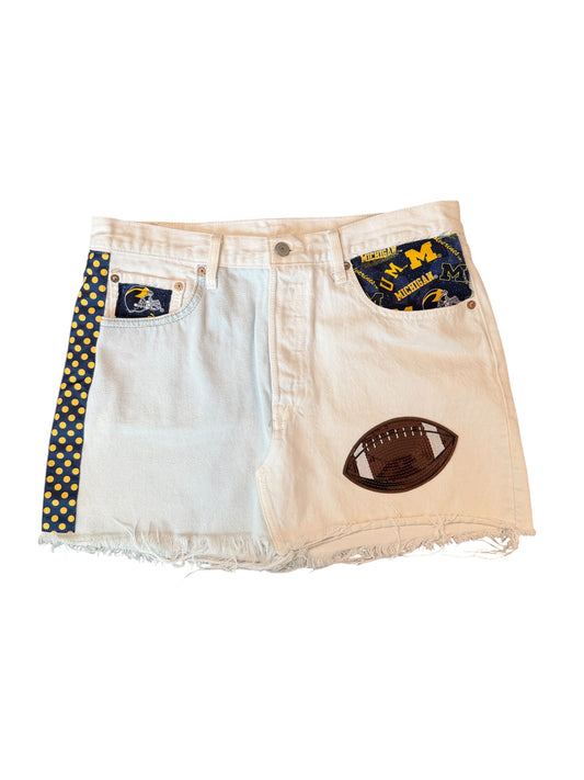 White denim shorts featuring a distressed frayed hem, a brown football patch on the right leg, a navy blue polka dot side stripe with gold dots, and a navy blue back pocket patch with the Michigan Wolverines 'M' logo.