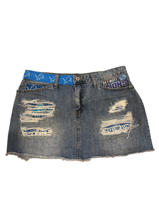 A distressed denim mini skirt with raw frayed edges, featuring ripped details and a blue patterned waistband with white text and graphics.
