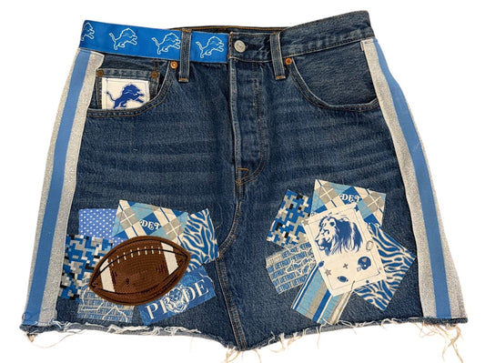 A pair of blue denim shorts featuring a patchwork design with various sports-themed prints, including a football patch, a blue and white patterned patch, and a small white patch with a blue logo. The shorts are laid flat on a white background.