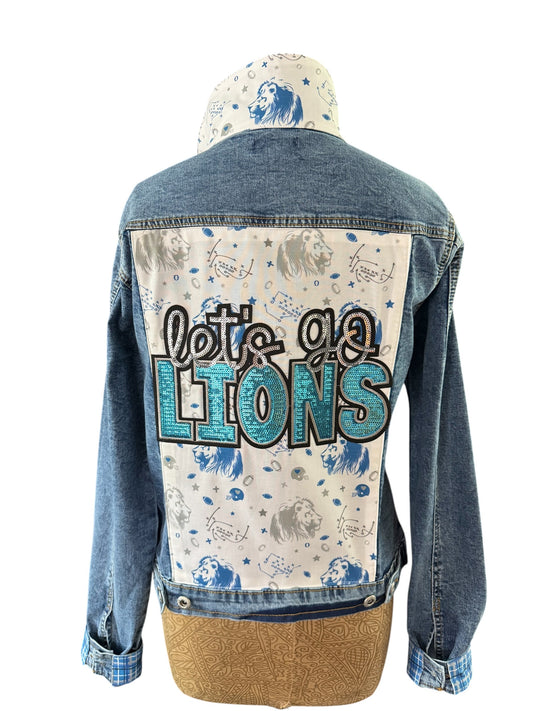 A denim jacket shown from the back on a dress form, featuring a light cream back panel with a blue lion print and the text 'let's go LIONS' in black and glittery blue.