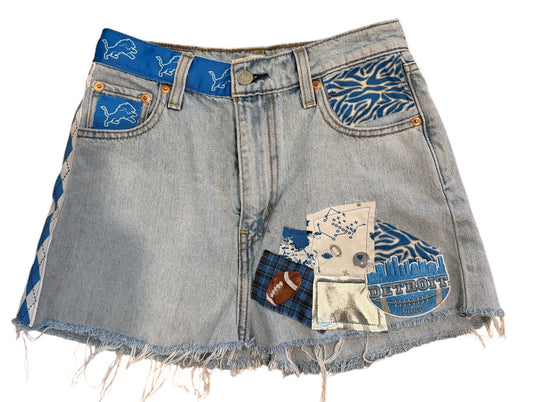 Light-wash denim shorts with frayed hems and decorative blue patchwork details on the back pockets, shown on a plain white background.