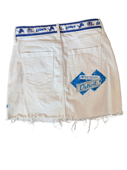 A pair of off-white denim shorts with a distressed frayed hem, featuring a blue and white Detroit Lions waistband and a blue diamond-shaped Detroit patch on the back pocket.