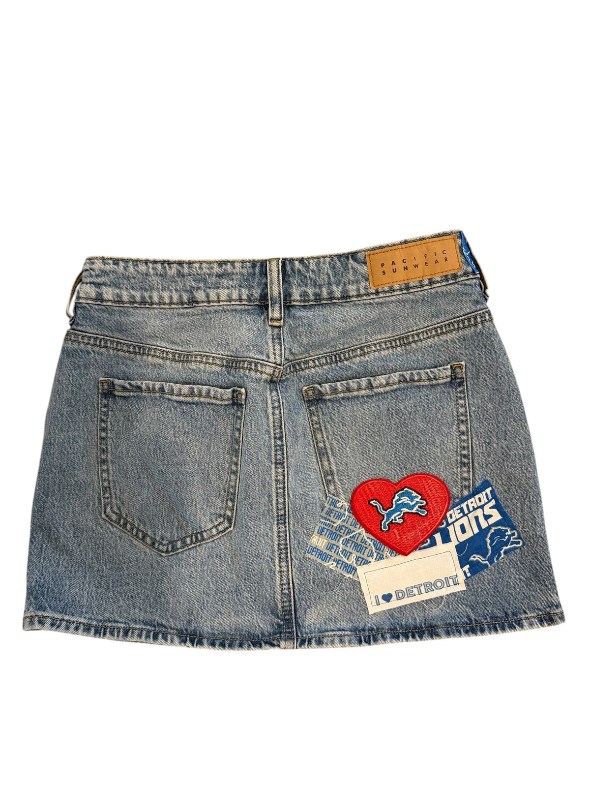 A light blue denim mini skirt shown from the back, featuring a Detroit Lions patch on the right pocket and a brown leather waistband patch.