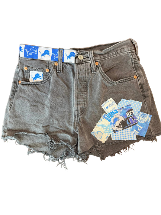 A pair of gray washed denim shorts with raw, frayed hems, laid flat on a dark wooden floor. The shorts feature decorative blue and white patches on the pocket trims and a small cluster of patches near the waistband.