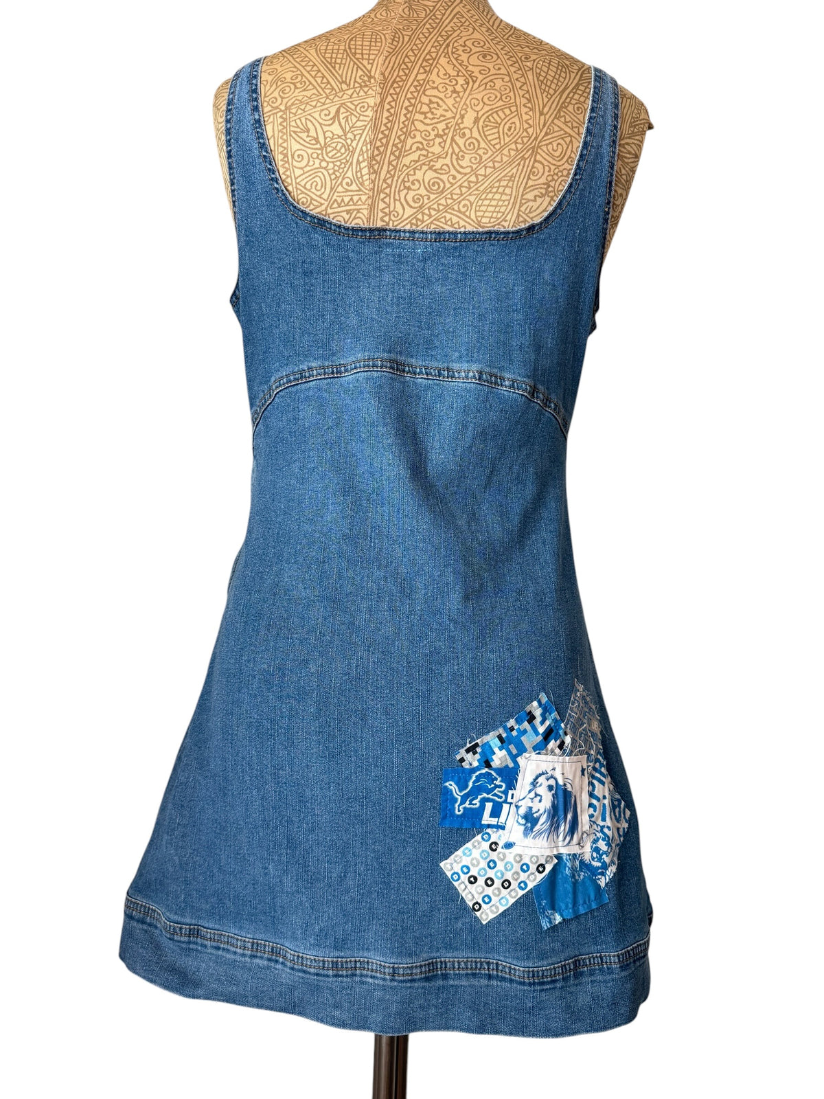 A sleeveless blue denim dress displayed on a mannequin torso against a white background. The dress features a scoop neckline, a curved hem, and a decorative blue and white patchwork applique on the lower right side.