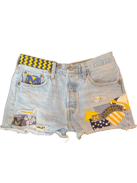 Light wash denim shorts with distressed details, frayed hems, and various Michigan State themed patches and a yellow-and-black chevron waistband.
