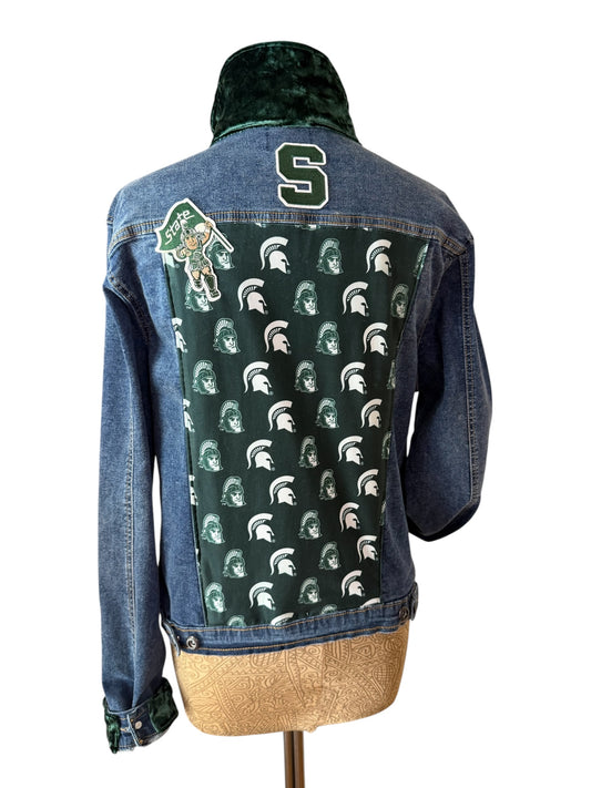 A denim jacket displayed on a dress form, showing the back view. The jacket features a dark green velvet collar, a green back panel covered in a repeating Michigan State Spartans helmet pattern, a green and white Spartan logo on the upper left, a large green number 5 on the upper back, and a gold glitter hem.