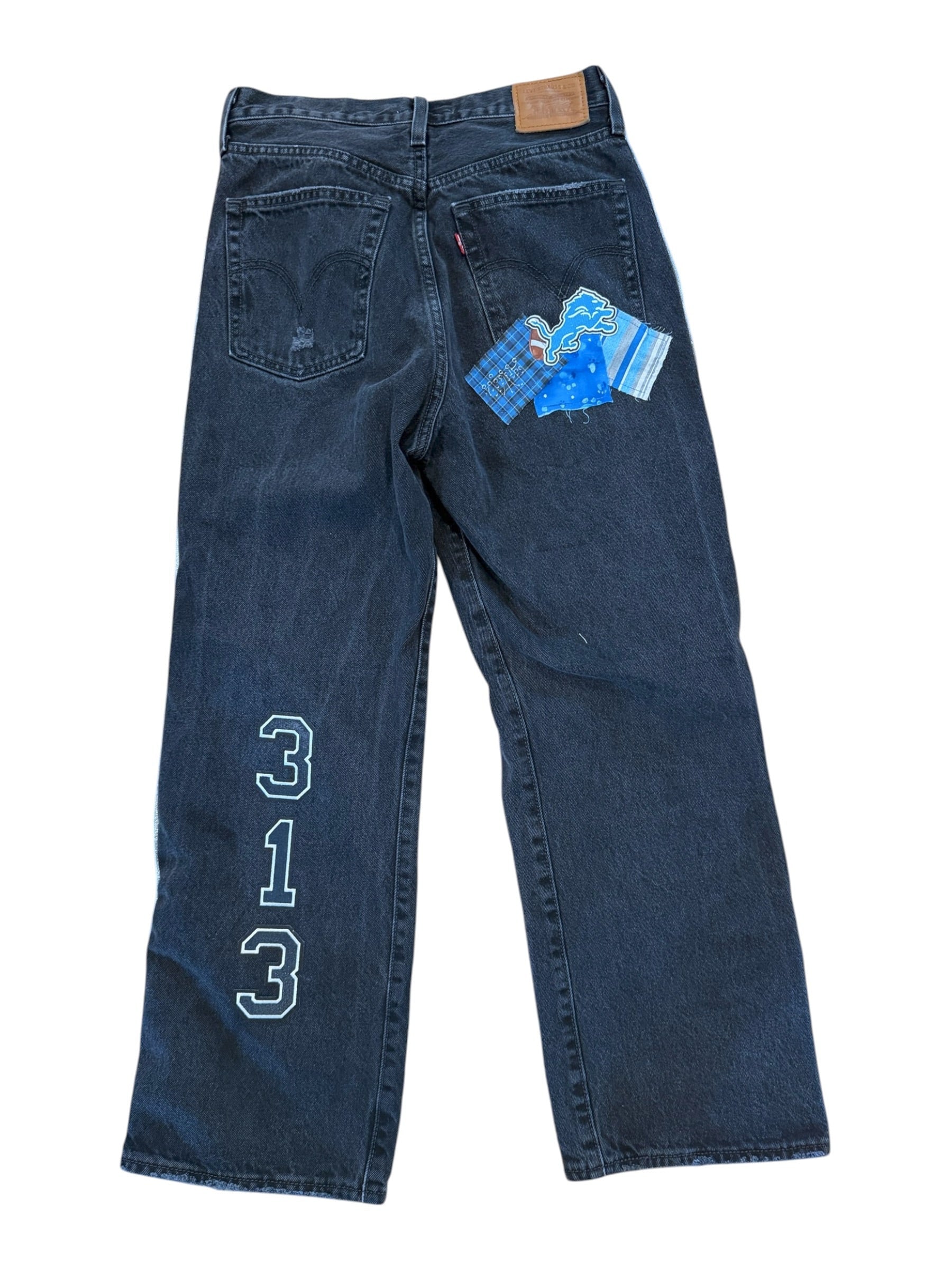 A pair of dark blue denim jeans laid flat on a white background, featuring a light blue embroidered geometric logo on the upper left leg and a blue patterned paper tag attached near the back pocket.