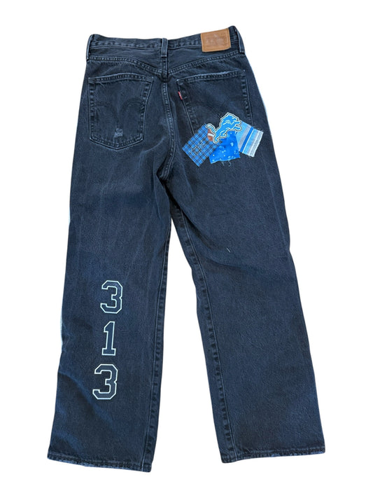 A pair of dark blue denim jeans laid flat on a white background, featuring a light blue embroidered geometric logo on the upper left leg and a blue patterned paper tag attached near the back pocket.