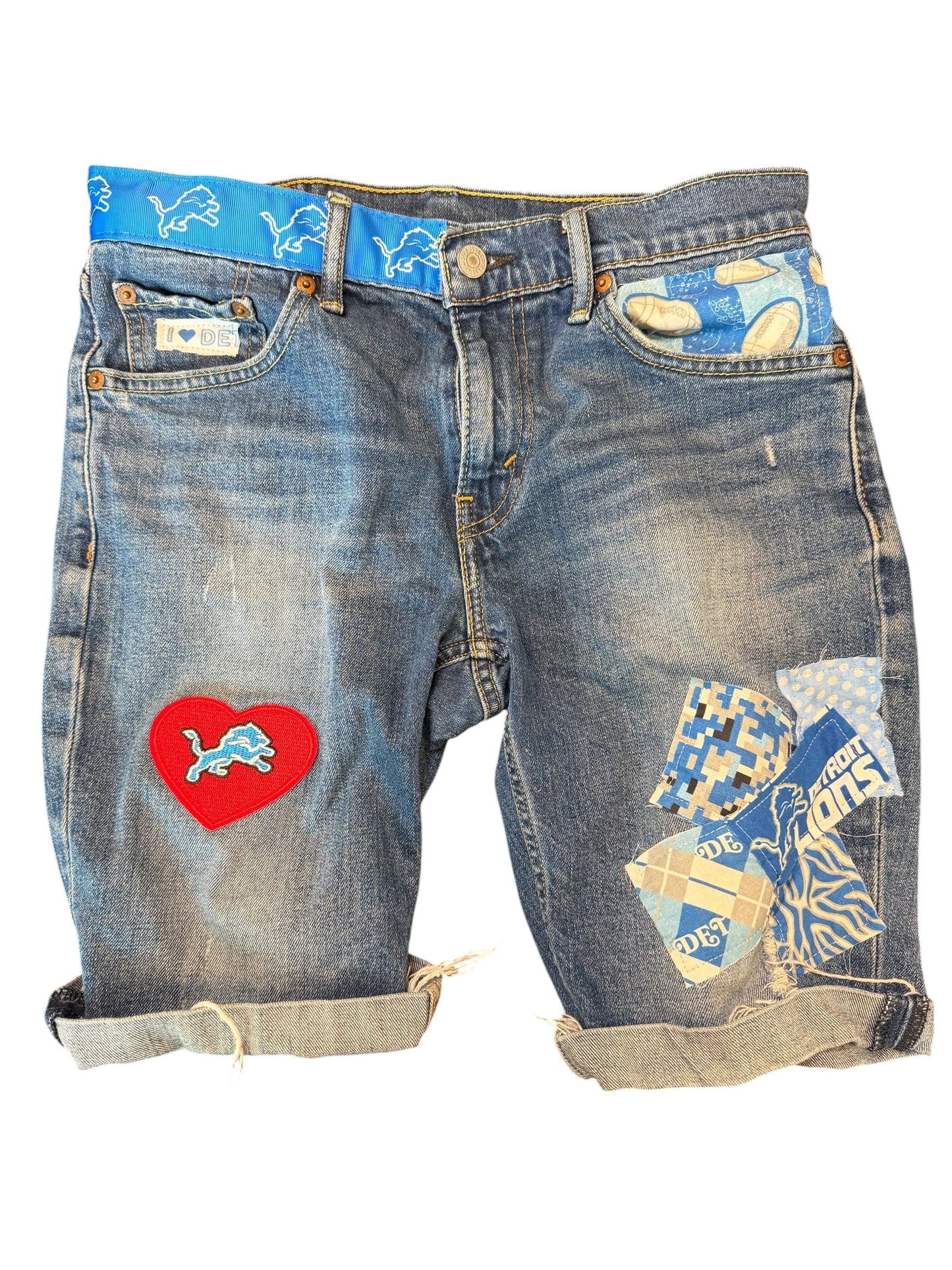A pair of distressed blue denim shorts with rolled cuffs, featuring a red heart patch with a blue lion logo on the left leg and a collage of blue, white, and tan patches on the right leg. The shorts are displayed flat against a white background.