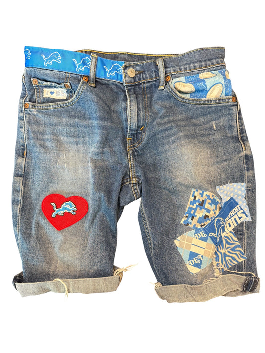 A pair of distressed blue denim shorts with rolled cuffs, featuring a red heart patch with a blue lion logo on the left leg and a collage of blue, white, and tan patches on the right leg. The shorts are displayed flat against a white background.