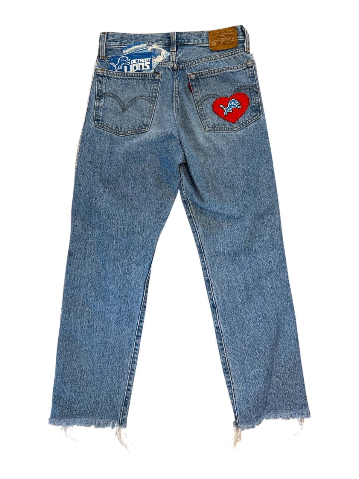 Light blue denim jeans laid flat on a white background, featuring a red heart-shaped patch on the right pocket and a blue and white 'Society' brand tag on the back pocket.