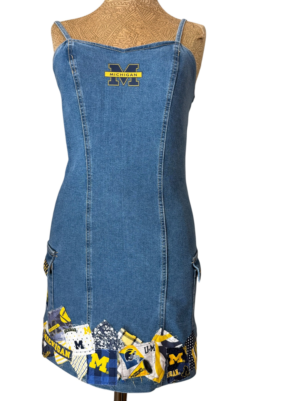 A blue denim dress displayed on a tan mannequin torso against a white background. The dress features a yellow and black Michigan 'M' logo on the upper chest and a patchwork hem with blue, yellow, black, and white collegiate patterns.