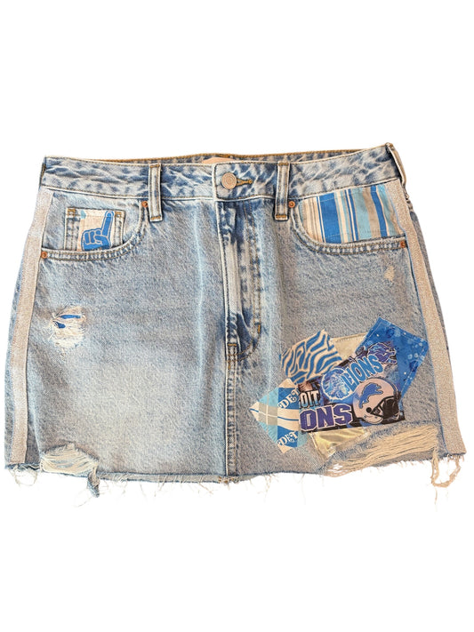Light wash blue denim mini skirt with distressed details, frayed hems, side stripes, and a blue graphic patch on the front pocket.