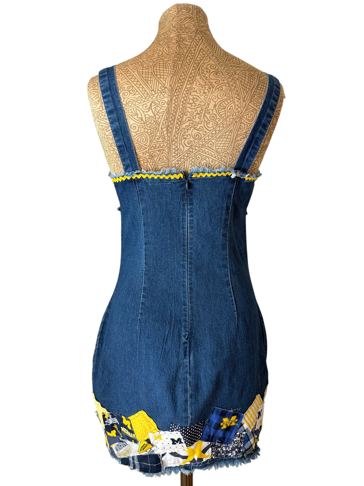 A denim corset-style dress or apron-style garment displayed on a tan mannequin torso against a white background. The garment has blue denim fabric, wide shoulder straps, a fitted waist, and decorative patchwork trim with yellow, white, and blue details at the hem and upper front edge.