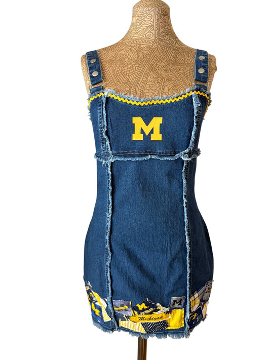 A blue denim pinafore-style dress displayed on a tan mannequin torso against a white background. The dress has a large yellow letter 'M' on the chest, yellow trim along the neckline, and a patchwork hem with yellow and black details.