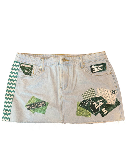 A pair of light-wash denim shorts featuring green and white chevron trim along the waistband and multiple green graphic patches with text and patterns on the front, displayed flat against a white background.