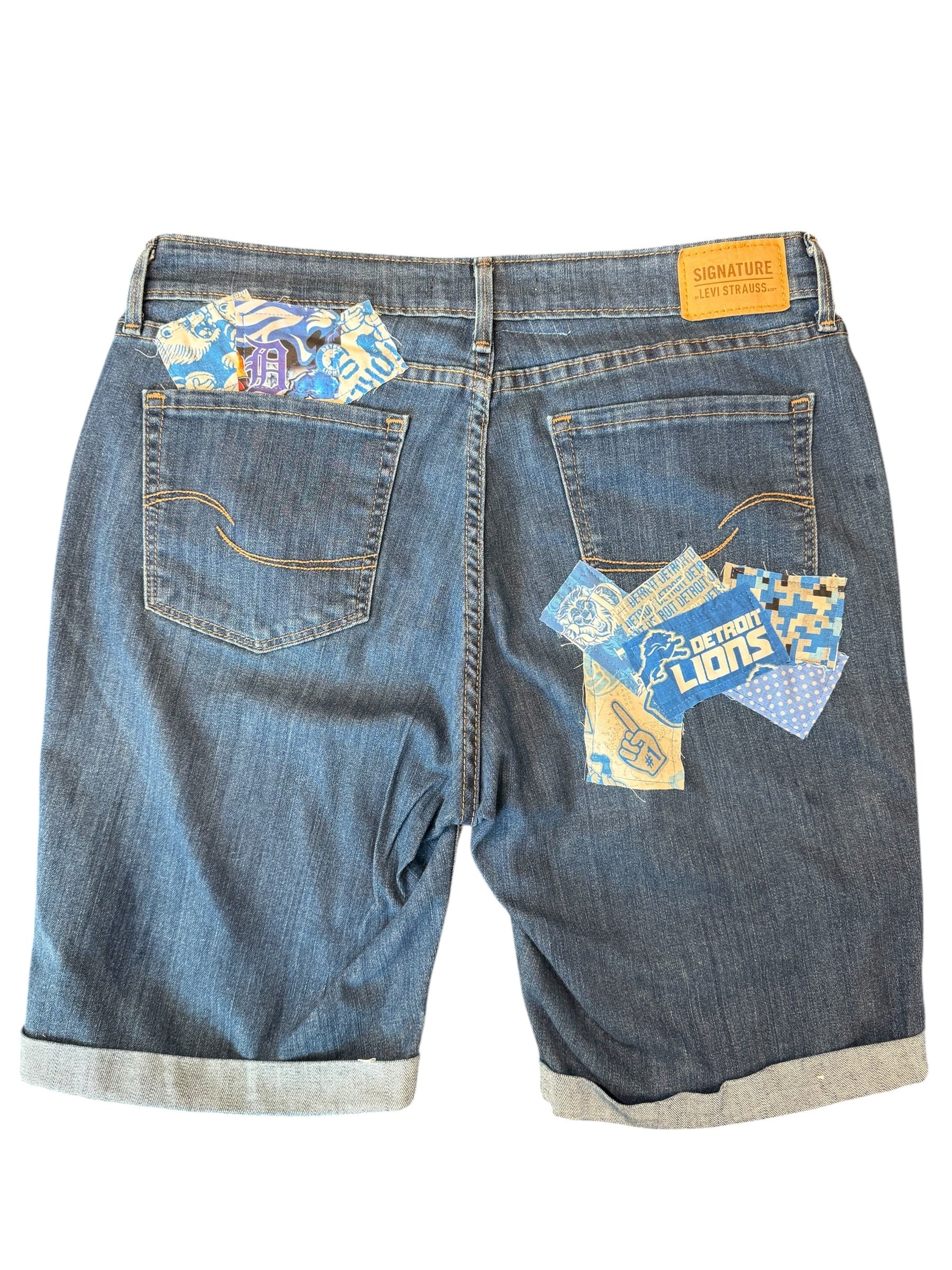 Back view of blue denim shorts with rolled cuffs, featuring decorative patchwork fabric inserts in the back pockets and a yellow leather brand patch at the waistband.