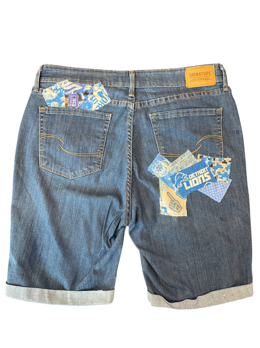 Back view of blue denim shorts with rolled cuffs, featuring decorative patchwork fabric inserts in the back pockets and a yellow leather brand patch at the waistband.