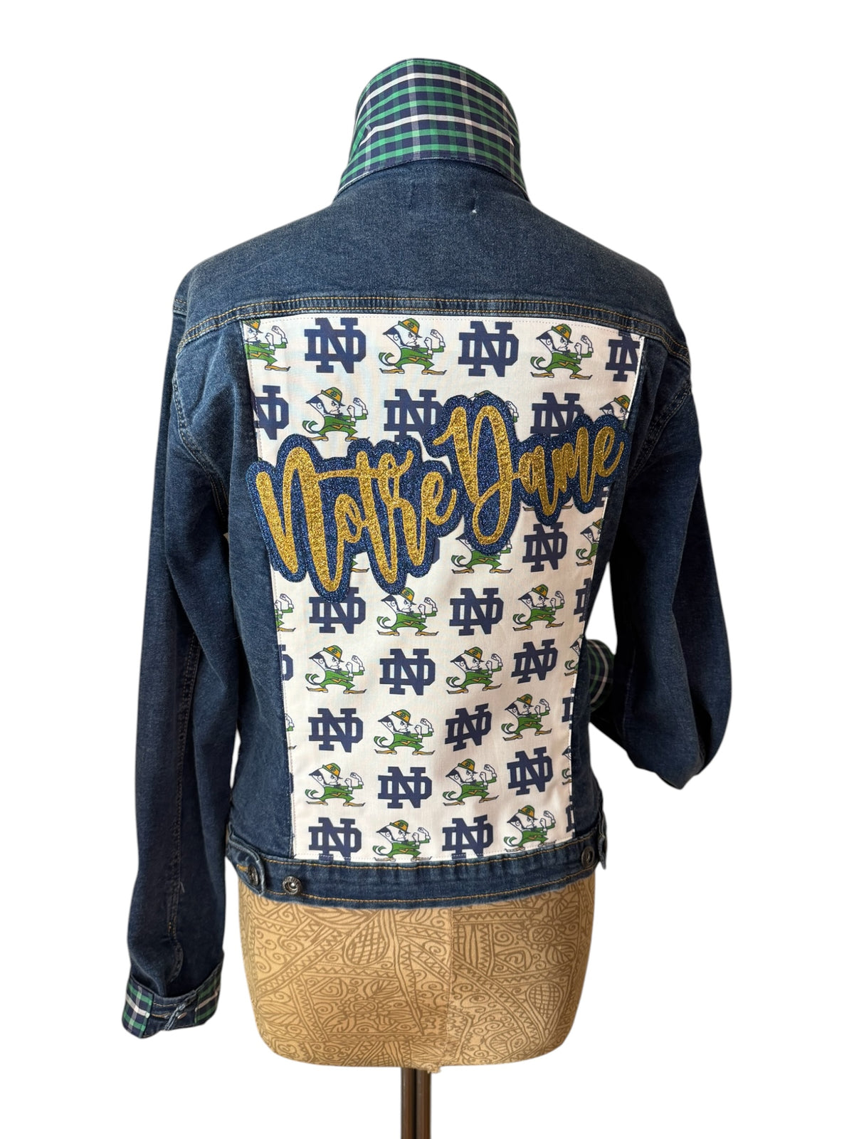 A denim jacket shown from the back on a dress form, featuring a large white back panel printed with Notre Dame logos and the word 'Notre Dame' in gold script, with a green plaid collar.