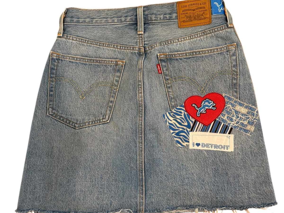 A light blue denim mini skirt with raw, frayed edges, featuring a patchwork design with a red heart patch, blue-and-white zebra print, and a brown leather patch on the back pocket area.