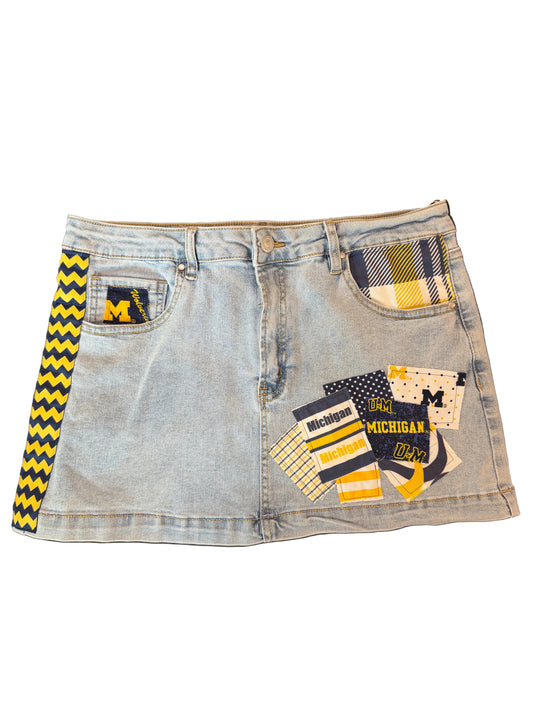A light wash denim mini skirt featuring a yellow and black chevron stripe down the left side, a plaid pocket patch, and a cluster of Michigan-themed patchwork fabric designs on the front right side.