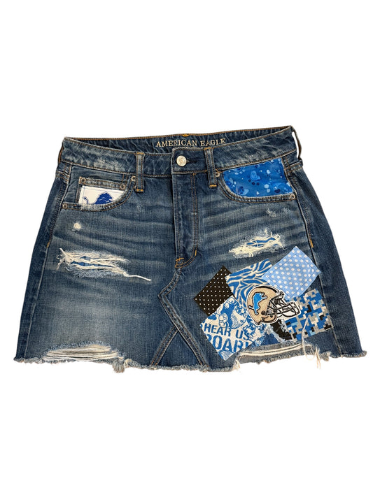 A distressed denim mini skirt with frayed hems, featuring a blue patterned back pocket patch, a football player patch, and 'AMERICAN EAGLE' text on the waistband, shown against a plain white background.