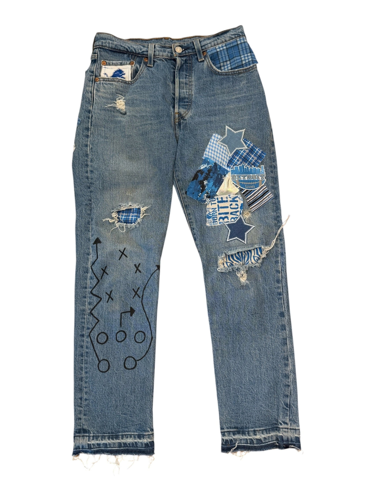 Light blue denim jeans with heavy distressing, ripped details, and a patchwork design on the right leg featuring blue plaid and striped fabrics. The left leg has black hand-drawn style graphics.