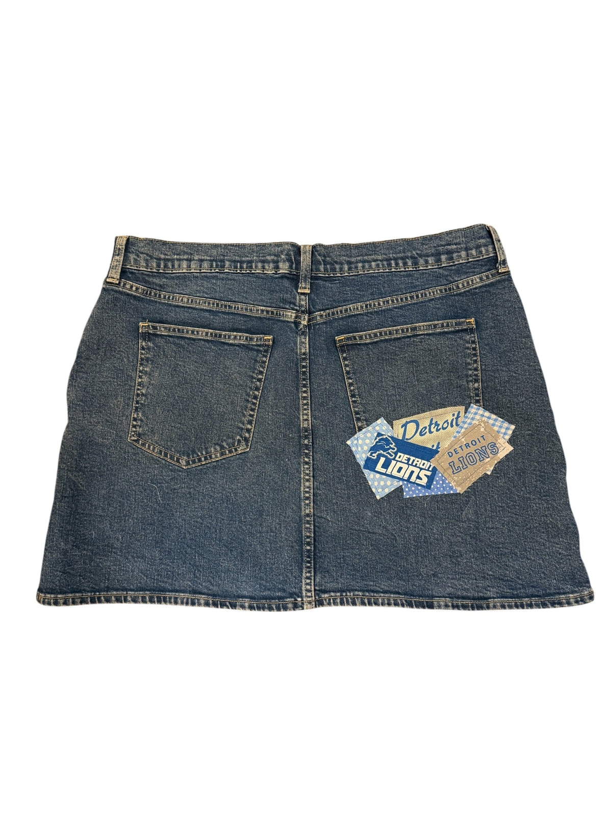 A pair of blue denim shorts with a distinctive patchwork design, featuring a large back pocket on the right side with a blue and white 'Suds of Choice' logo patch, and a smaller tan patch on the left side. The shorts are laid flat against a white background.