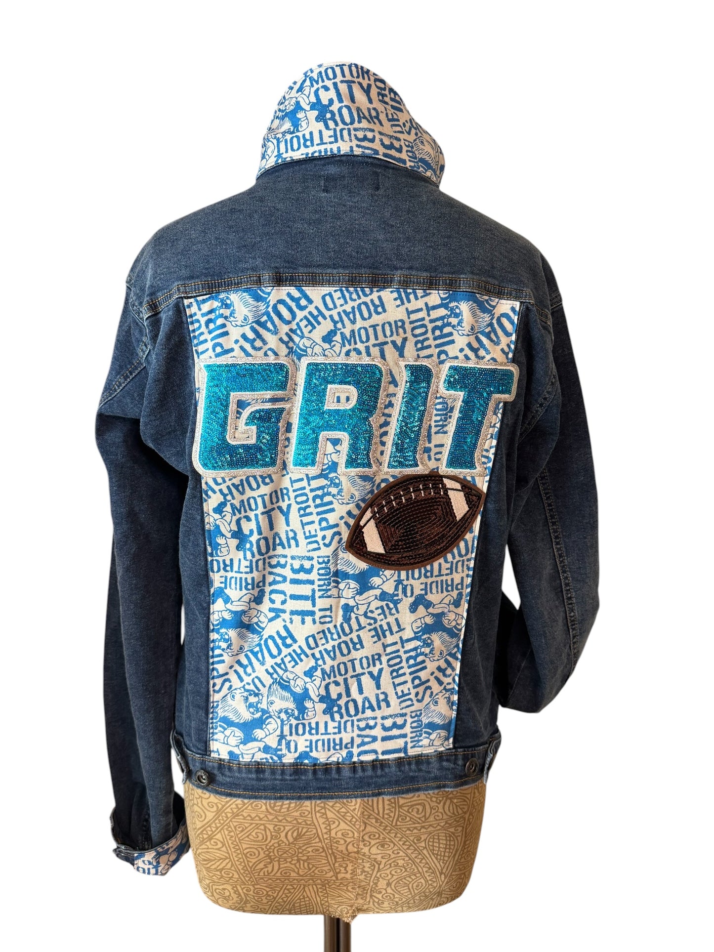 A blue denim hooded jacket shown from the back on a dress form, featuring a large white patch with blue text reading 'GRIT', a blue hood lining with text, and a brown football patch on the back.