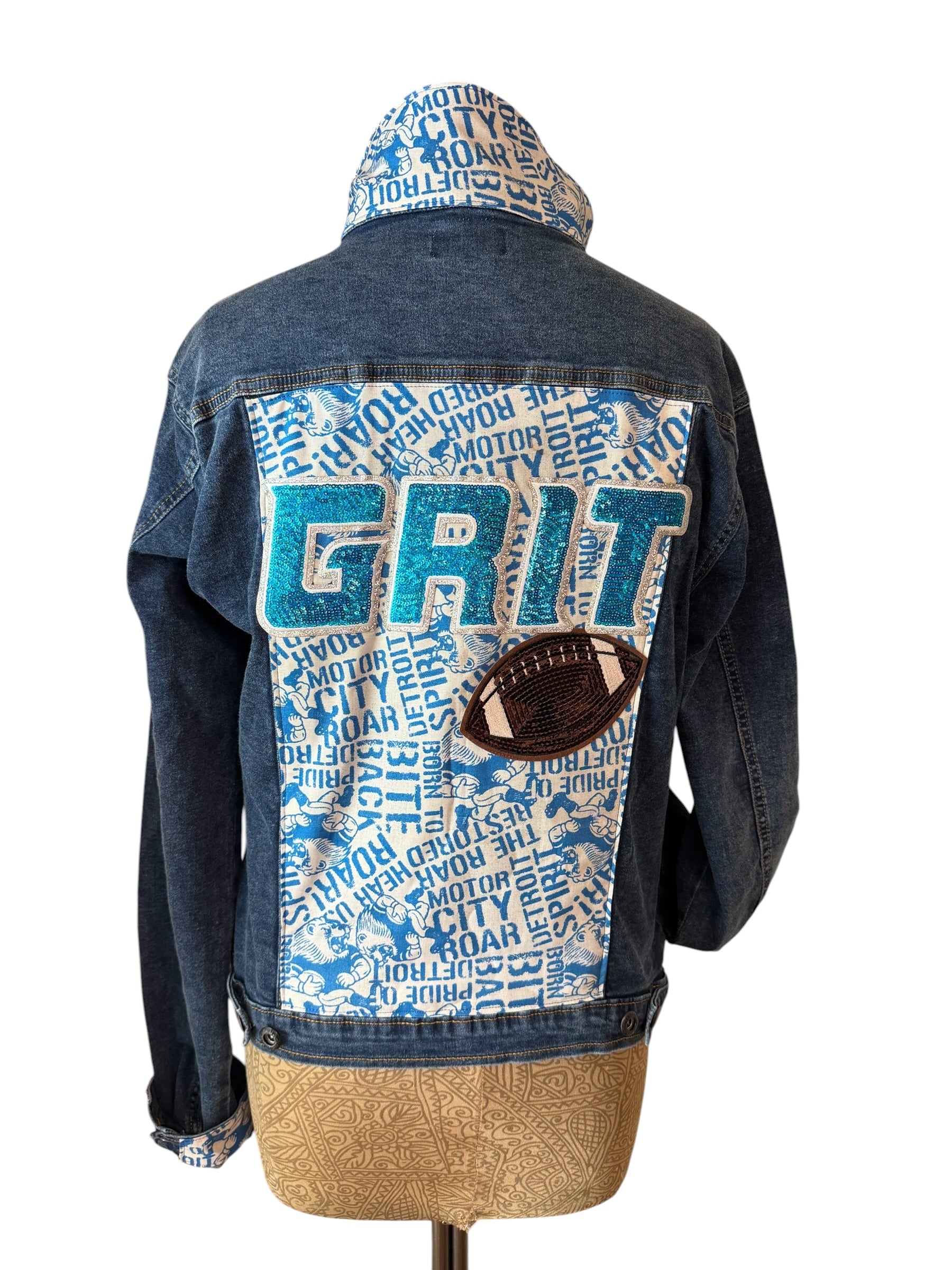 A blue denim hooded jacket shown from the back on a dress form, featuring a large white patch with blue text reading 'GRIT', a blue hood lining with text, and a brown football patch on the back.