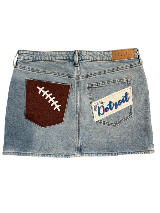 Light wash blue denim mini skirt shown from the back, featuring a brown back pocket with a white football patch and a white back pocket with a Detroit patch.