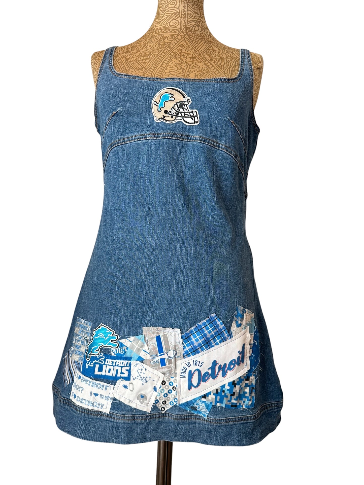 A blue denim apron-style dress displayed on a mannequin torso against a white background. The dress features a Detroit Lions helmet patch near the chest and a decorative patchwork collage at the bottom with 'Detroit Lions' text and team graphics.