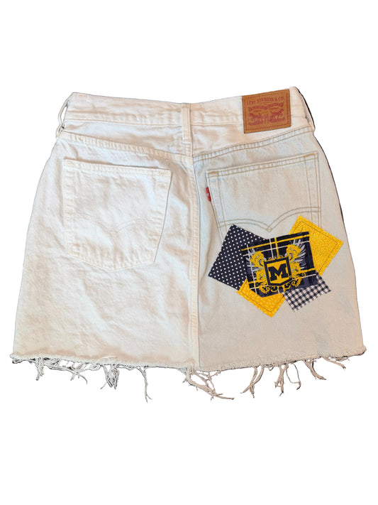Off-white denim shorts with a distressed frayed hem, featuring a patchwork design on the back pocket with yellow, navy, and black patterns, and a brown leather patch on the waistband.