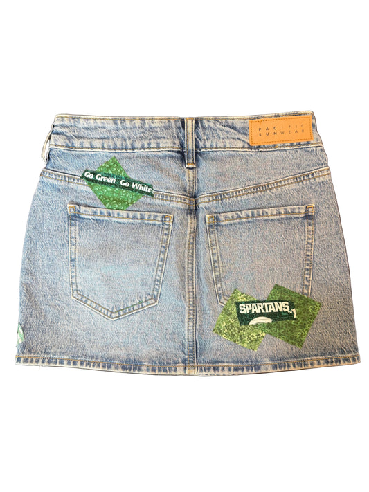 A light wash blue denim mini skirt shown from the back, featuring two green glittery 'SPARTANS' patches on the back pockets and a tan leather brand patch at the waistband.