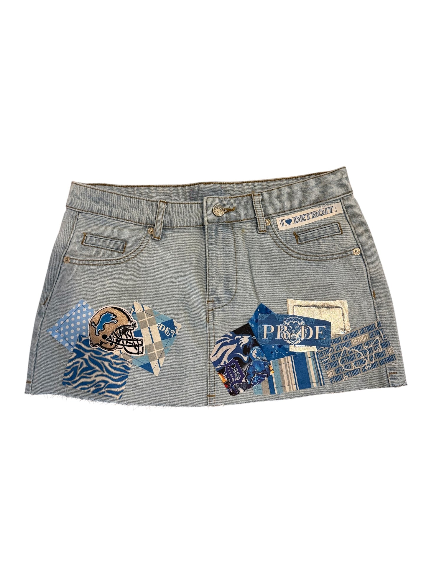 A light blue denim mini skirt with distressed details, featuring custom patches on the front pockets. The patches include a Detroit Lions helmet, blue patterned fabrics, and a patch with the word 'PROUD'.