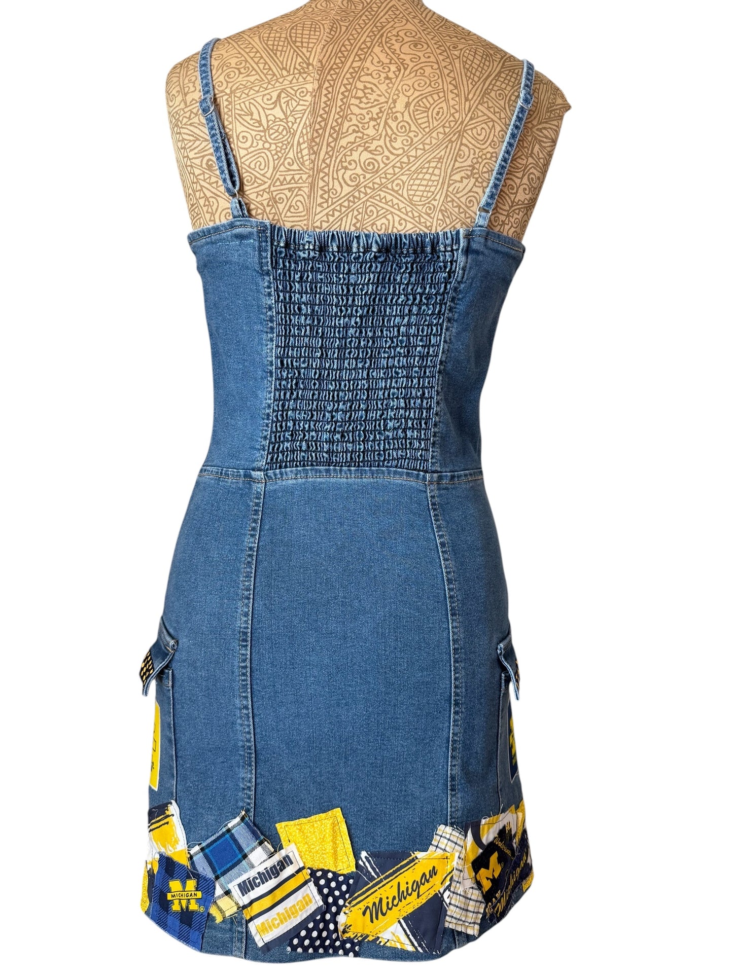 A blue denim dress displayed on a tan mannequin torso against a white background. The dress features a smocked bodice, thin shoulder straps, and a patchwork hem with yellow, black, and white collegiate-style fabric panels.