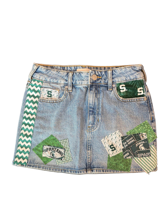 A light blue denim mini skirt featuring green and white patchwork details, including a green and white chevron stripe on the left side, a green patch with a white 'S' on the right pocket, and other decorative patches on the front.