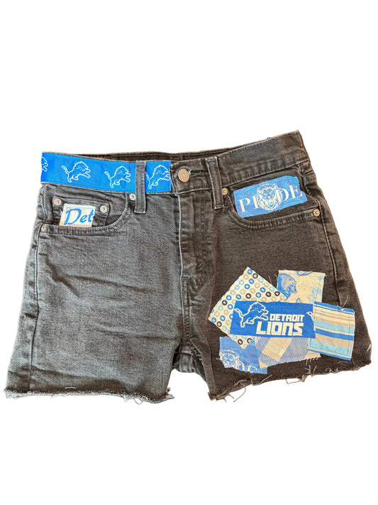 A pair of dark gray denim shorts with raw, frayed hems, featuring multiple blue and white 'Detroit Lions' team patches attached to the front pockets, laid flat on a wooden surface.