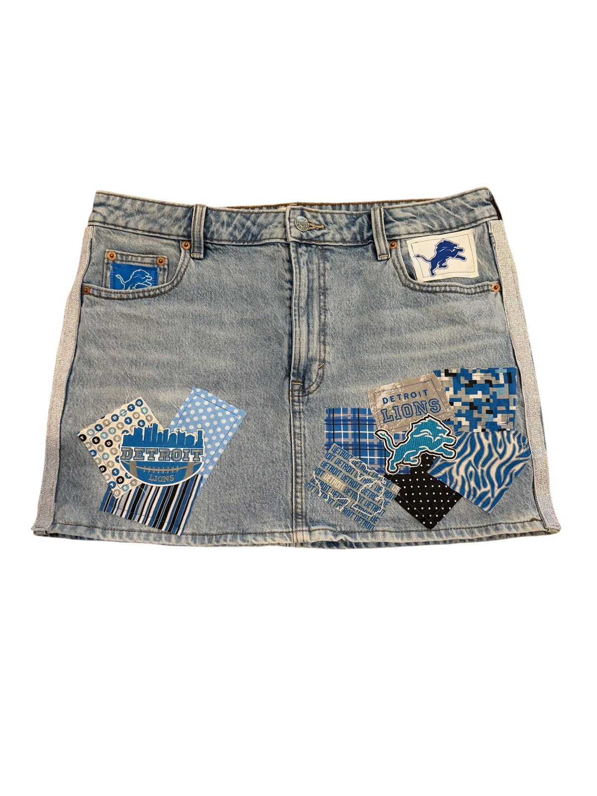 A light blue denim mini skirt featuring Detroit Lions themed patches, including a lion logo patch on the right pocket and a patterned patch on the left pocket.