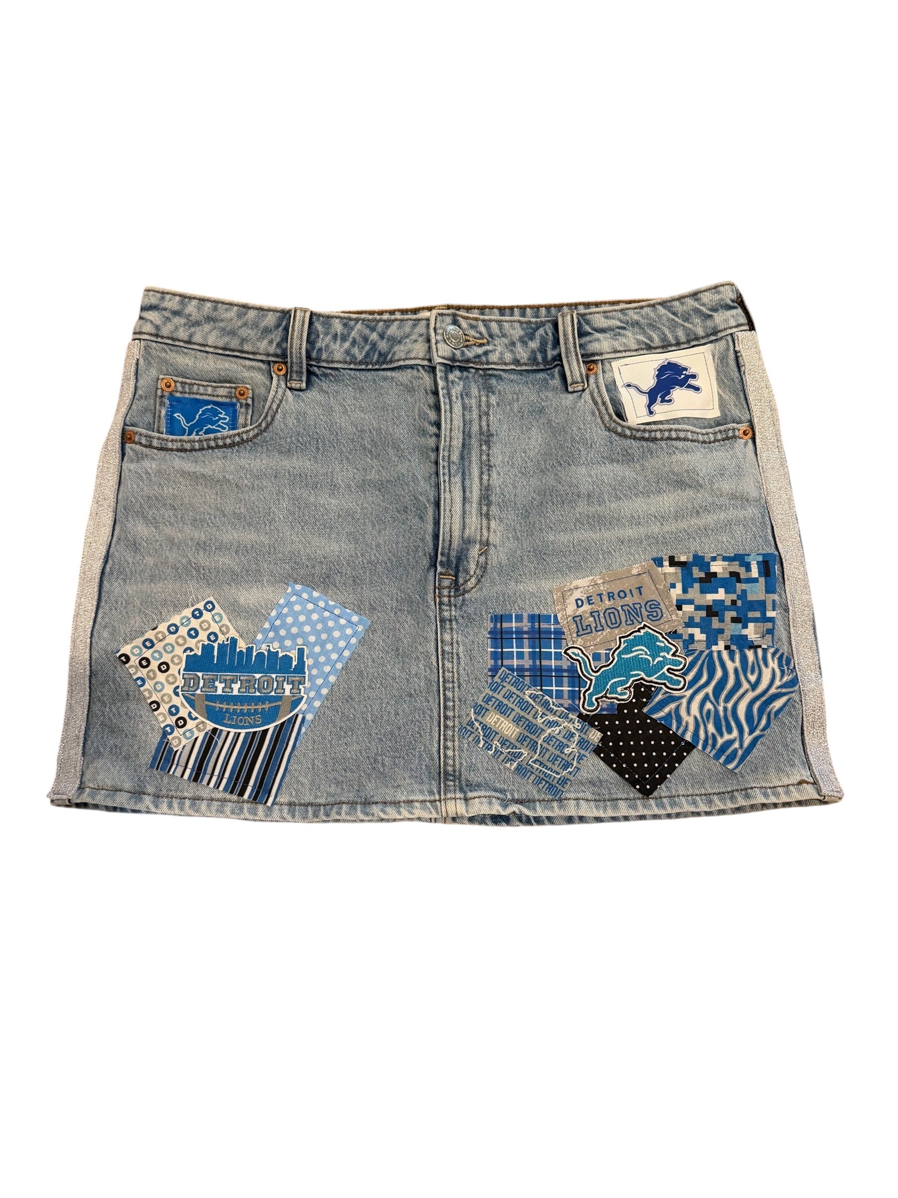 A light blue denim mini skirt featuring Detroit Lions themed patches, including a lion logo patch on the right pocket and a patterned patch on the left pocket.