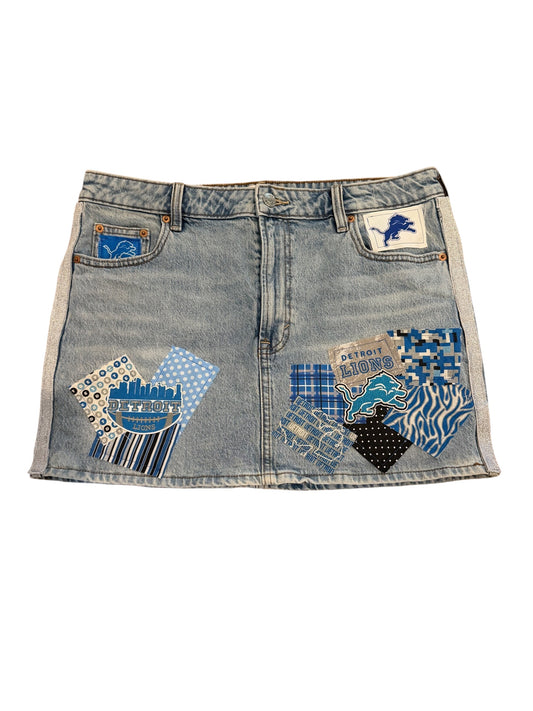 A light blue denim mini skirt featuring Detroit Lions themed patches, including a lion logo patch on the right pocket and a patterned patch on the left pocket.