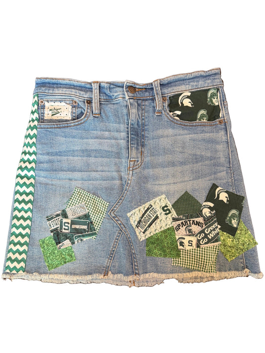 A light blue denim mini skirt with a raw, frayed hem and decorated with green and black patchwork fabric details, including a green and white chevron stripe on the side.