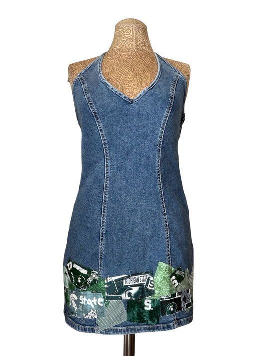 A sleeveless blue denim tunic dress displayed on a tan mannequin torso against a white background. The dress features a V-neckline, front seam detailing, and a green Michigan State themed patchwork collage at the hem.